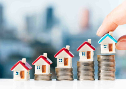 Concept for property ladder, mortgage and real estate investment. Man's hand putting blue house model on top of coins stack with city backgrounds.