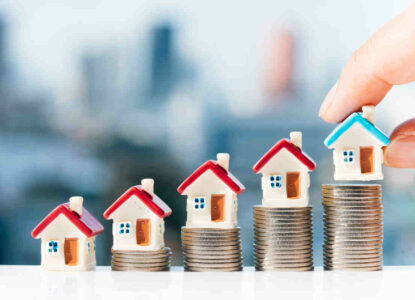 Concept for property ladder, mortgage and real estate investment. Man's hand putting blue house model on top of coins stack with city backgrounds.